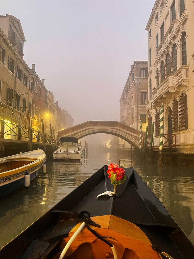View from the bow of a gondola gliding down a misty canal towards a small arched bridge, moored boats either side
